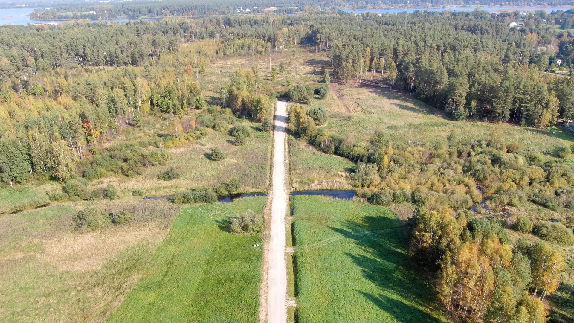 Prieka iela no augšas rudenī: grants iela starp nopļautiem zemesgabaliem, pār meliorācijas grāvi — pārbrauktuve, pie horizonta Daugava